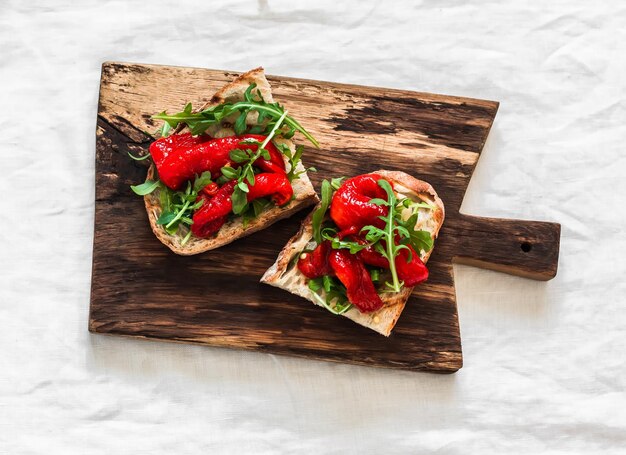 Photo delicious snack appetizer tapas grilled bread with cream cheese arugula and baked sweet peppers on a wooden cutting board on a light background