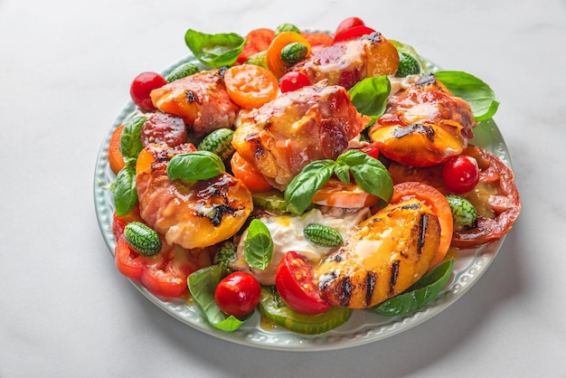 Delicious summer salad with grilled peaches tomatoes prosciutto and cheese in a plate on white table Close up