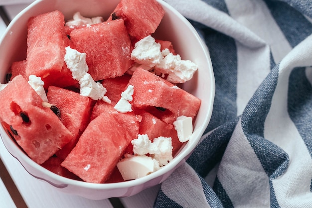 Delicious summer salad with slices of watermelon and feta cheese on white bowl with a striped towel on a wooden white surface