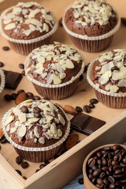 Delicious, sweet chocolate muffins, with almond petals in a wooden tray next to grains of coffee on a blue wooden table.