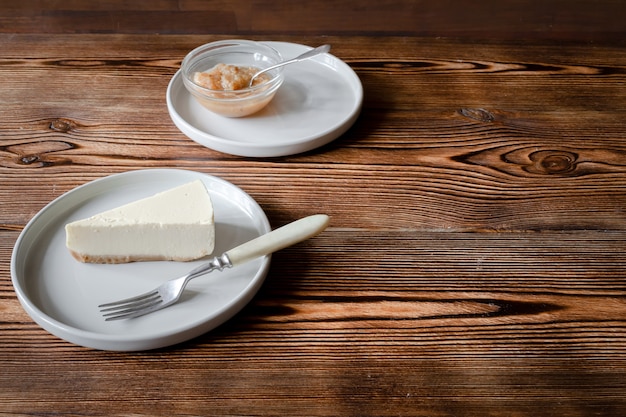 Delicious sweet dessert classical cheesecake New York slice on wooden rustic background. Slice of tasty cake on ceramic plate served with dessert fork and apple jam.