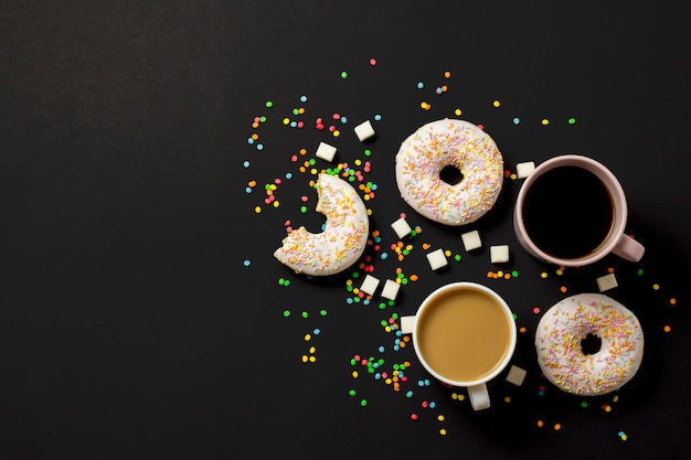 Delicious, sweet, fresh donuts, multicolored decorative candy, a cup of coffee on a black background. Concept of breakfast, fast food, coffee shop, bakery. Flat lay, top view.