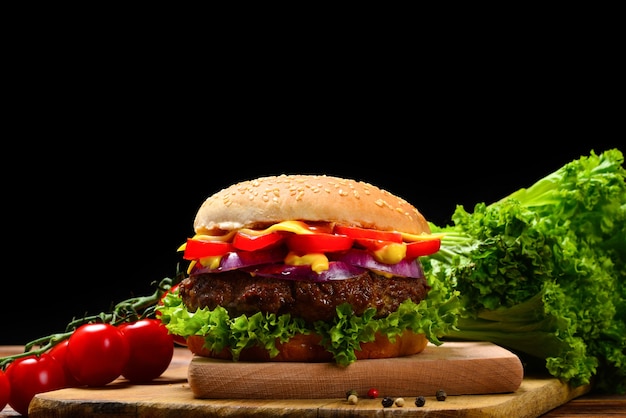 Delicious tasty burger with beef, tomatoes, salad and sauce on wooden table and black background.  Space for text or design.