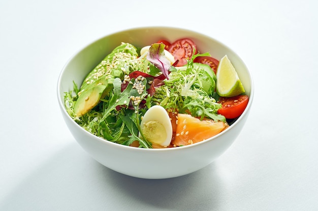 Delicious veggie bowl with salmon, avocado, cucumber, cherry tomatoes and mix salad in a white plate. Isolated on white surface.