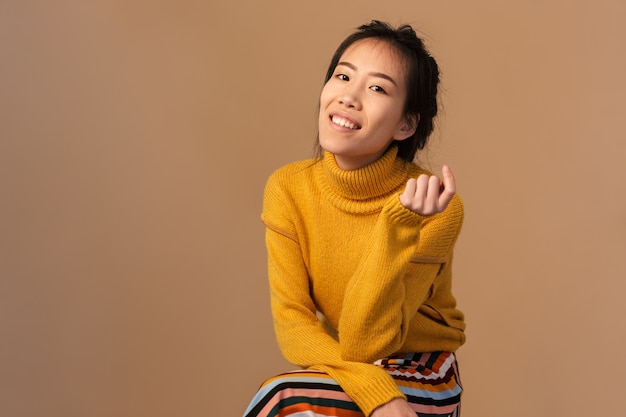 delighted asian woman wearing sweater smiling while sitting in studio isolated over beige wall