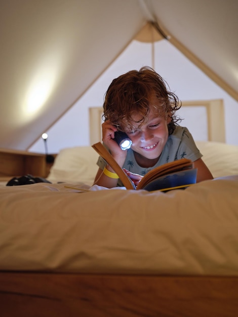 Delighted boy with shining flashlight reading interesting book with smile on comfortable bed on mansard at home on evening time