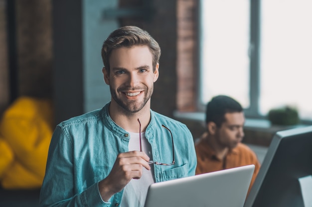 Delighted good looking man standing with his glasses in the hand while reading from the laptop screen