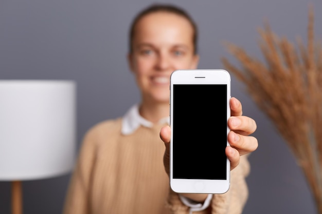 Delighted smiling woman wearing beige sweater posing in stylish room holding cell phone looking at camera and showing phone display copy space for advertisement or commercial text