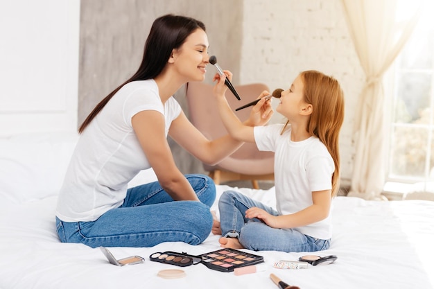 Delighted woman and her pleasant child powdering each other nose while sitting face to face and keeping legs crossed