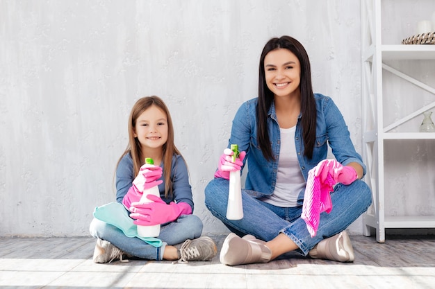 Delighted woman sitting cross legged with her pleasant daughter while wearing pink gloves and using air disinfectant