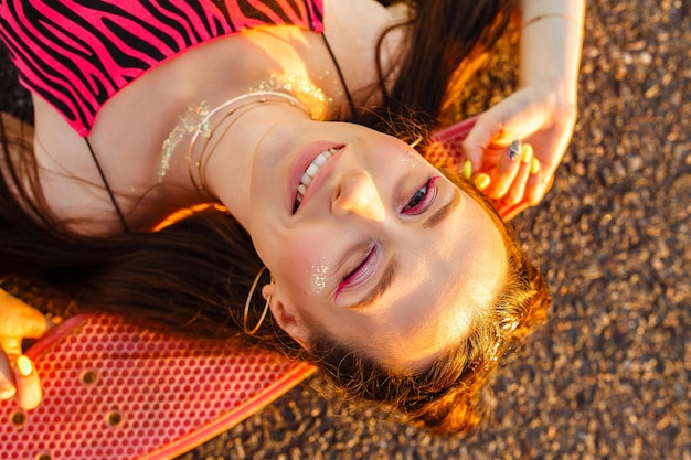 delighted young hipster woman lying on skateboard or penny board on road and looking at camera