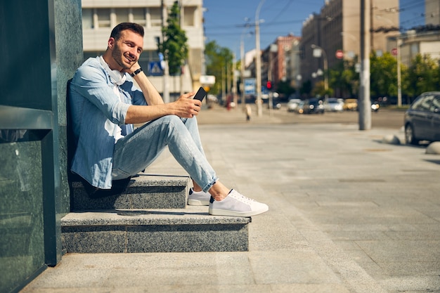 Delighted young man expressing positivity, holding telephone in right hand while waiting outdoors