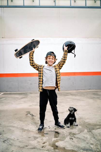 Delightful boy standing against concrete street wall with dog and looking at camera while raising skateboard with helmet