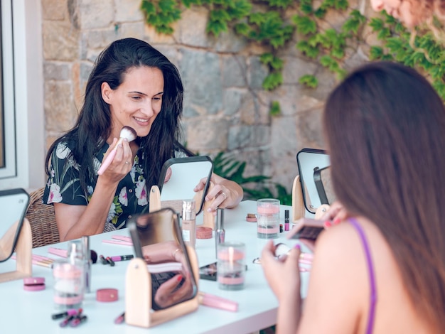 Delightful brunette and anonymous blurred lady sitting together at table with cosmetics and applying makeup against building wall in daylight