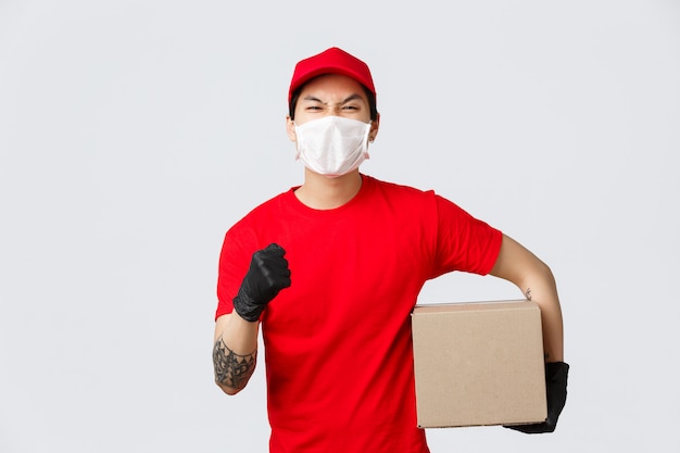 Deliver your goods no matter what. Confident asian delivery guy in red uniform cap and t-shirt, raise hand encourage place your order, holding box pachage, wear medical mask and gloves