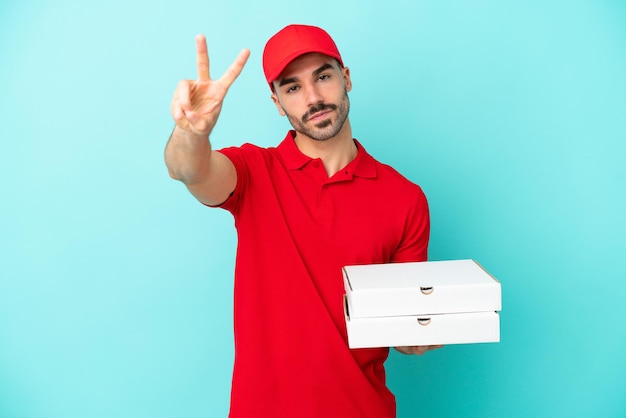 Delivery caucasian man picking up pizza boxes isolated on blue background smiling and showing victory sign