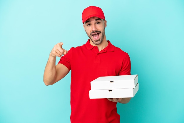 Delivery caucasian man picking up pizza boxes isolated on blue background surprised and pointing front