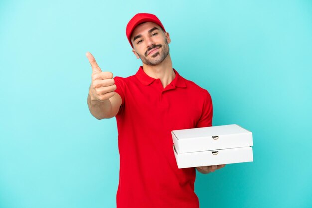 Delivery caucasian man picking up pizza boxes isolated on blue background with thumbs up because something good has happened