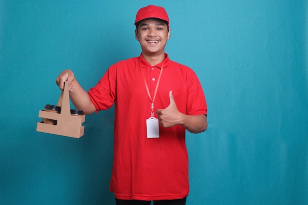 Delivery guy in red uniform holding takeaway mug drink in hand and giving thumb up in blue studio ba