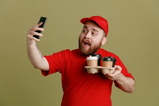 Delivery man employee in red cap blank tshirt uniform holding coffee cups doing selfie using smartphone smiling cheerfully standing over green background