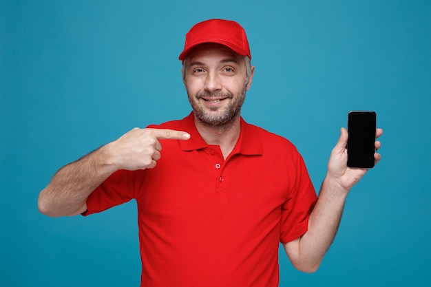 Delivery man employee in red cap blank tshirt uniform holding smartphone pointing with index finger at it looking at camera smiling standing over blue background