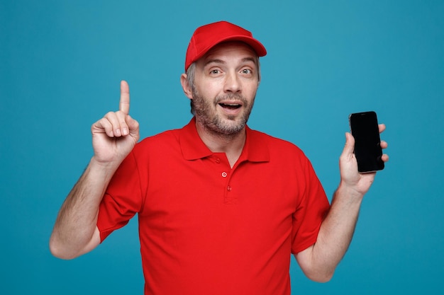 Delivery man employee in red cap blank tshirt uniform holding smartphone pointing with index finger up looking at camera smiling standing over blue background