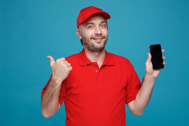 Delivery man employee in red cap blank tshirt uniform holding smartphone pointing with thumb to the side looking at camera smiling standing over blue background