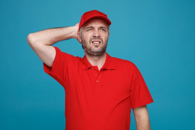 Delivery man employee in red cap blank tshirt uniform looking aside confused holding hand on his head for mistake standing over blue background