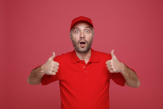 Delivery man employee in red cap blank tshirt uniform looking at camera being surprised showing thumbs up standing over red background