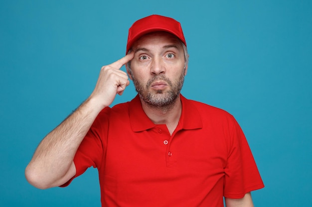 Delivery man employee in red cap blank tshirt uniform looking at camera confused pointing with index finger at his temple standing over blue background