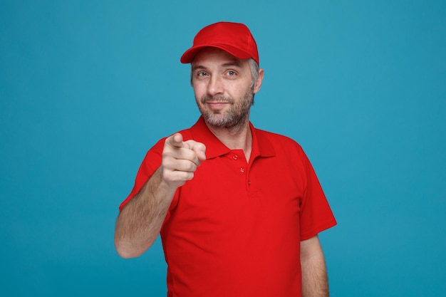 Delivery man employee in red cap blank tshirt uniform looking at camera happy and positive pointing with index finger at camera smiling standing over blue background