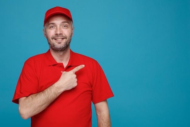 Delivery man employee in red cap blank tshirt uniform looking at camera happy and positive smiling cheerfully pointing with index finger to the side standing over blue background