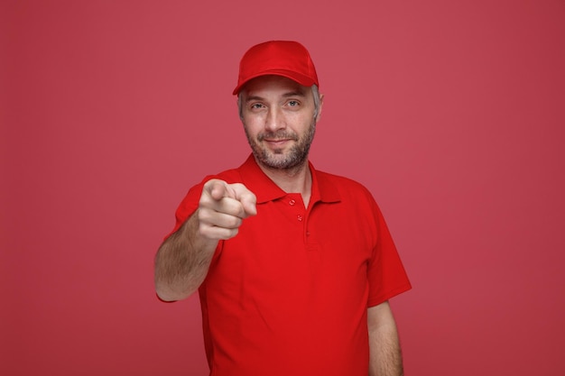 Delivery man employee in red cap blank tshirt uniform smiling friendly pointing with index finger at camera standing over red background