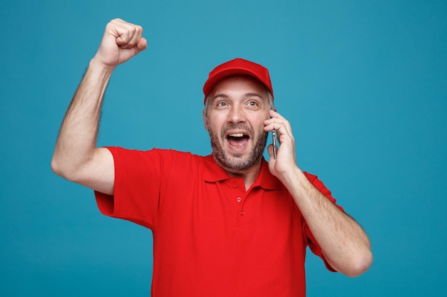 Delivery man employee in red cap blank tshirt uniform talking on mobile phone crazy happy and excited raising clenched fist standing over blue background