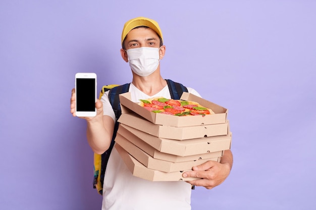 Delivery man employee wearing protective mask holding food order pizza boxes and showing mobile phone with blank screen isolated on purple background copy space for advertisement