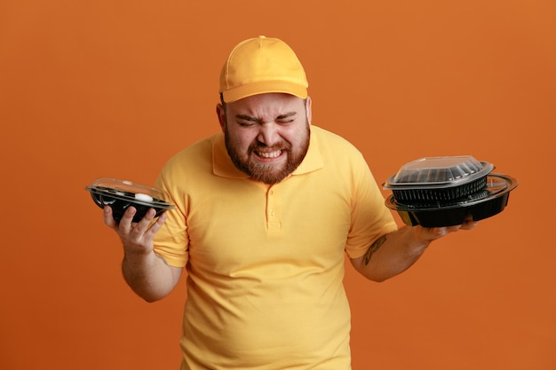 Delivery man employee in yellow cap blank tshirt uniform holding food containers angry and annoyed standing over orange background