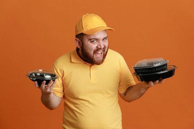 Delivery man employee in yellow cap blank tshirt uniform holding food containers looking at camera crazy happy and cheerful smiling standing over orange background