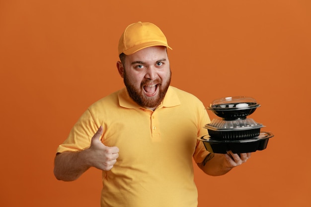 Delivery man employee in yellow cap blank tshirt uniform holding food containers looking at camera happy and excited smiling broadly showing thumb up standing over orange background