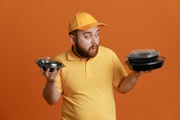 Delivery man employee in yellow cap blank tshirt uniform holding food containers looking confused having doubts standing over orange background