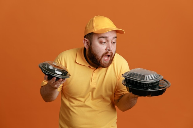 Delivery man employee in yellow cap blank tshirt uniform holding food containers looking surprised and confused standing over orange background