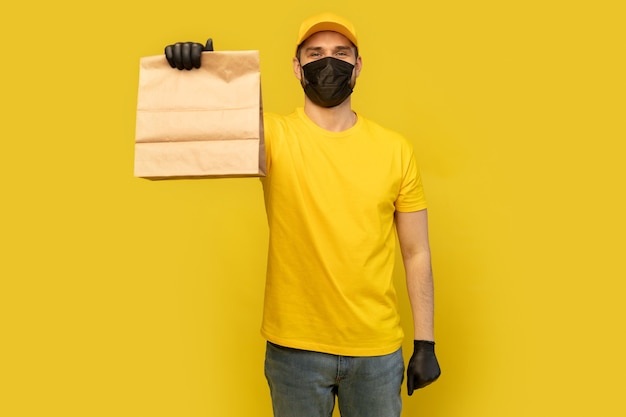 Delivery man employee in yellow cap tshirt uniform mask glove hold craft paper packet with food isolated