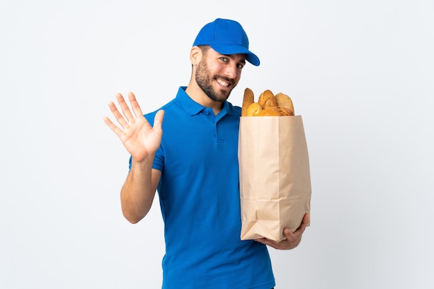 Delivery man holding a bag full of breads isolated on white background saluting with hand with happy expression