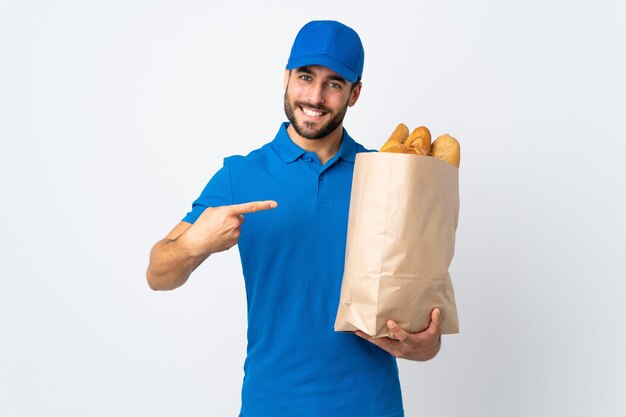 Delivery man holding a bag full of breads on white wall and pointing it