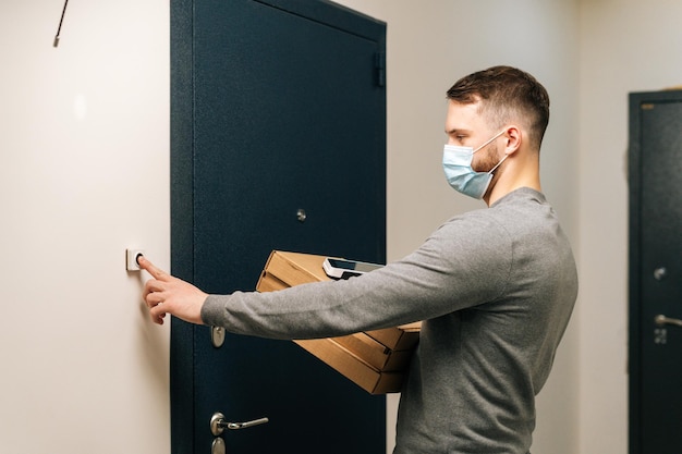 Delivery man in medical mask ringing doorbell of customer apartment holding in hand carton boxes with pizza and contactless payment POS terminal