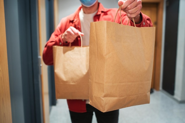 Delivery man in protective mask holding paper bag with food in the entrance. The courier gives the box with fresh vegetables and fruits to the customer