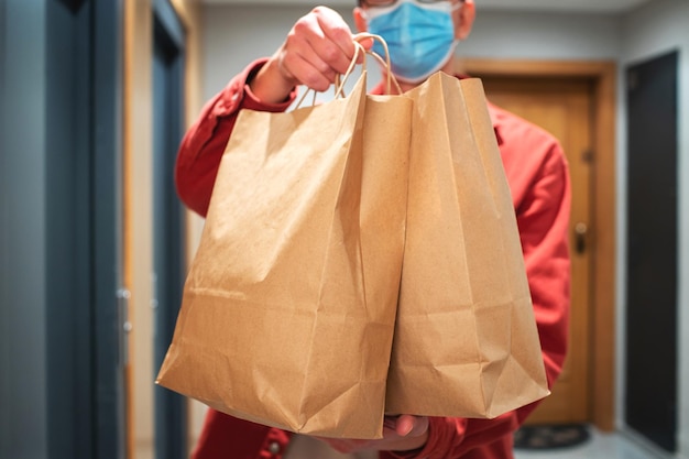 Delivery man in protective mask holding paper bag with food in the entrance. The courier gives the box with fresh vegetables and fruits to the customer