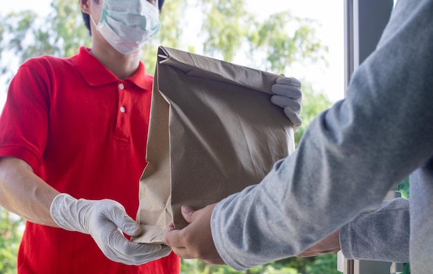 Delivery man in red tshirt uniform face mask gloves hold paper bag Customer receiving order from courier at home Service during quarantine 2019ncov coronavirus concept