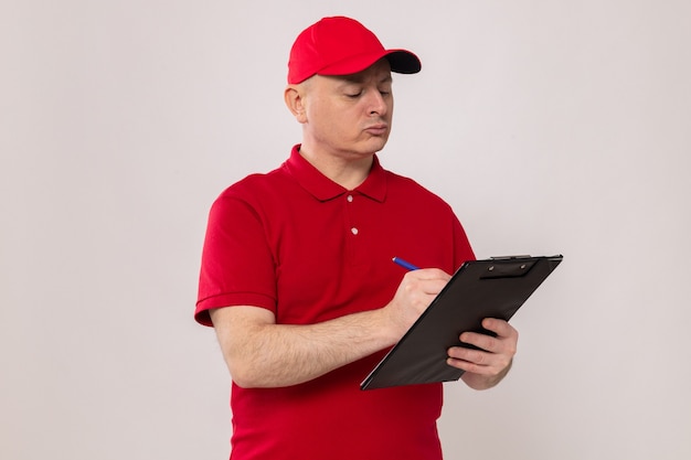 Delivery man in red uniform and cap holding clipboard and pen making notes with serious face