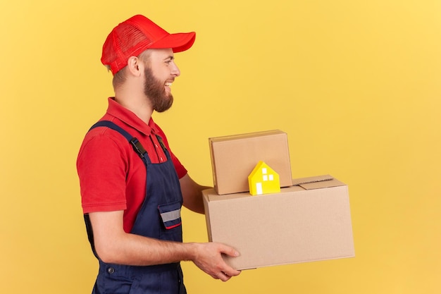 Delivery man in uniform and red cap holding cardboard boxes and paper house doortodoor delivery