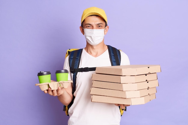 Delivery man wearing t shirt and cap holding pizza boxes and takeaway coffee in hands delivering food during flu epidemic bringing orders in time posing isolated over purple background